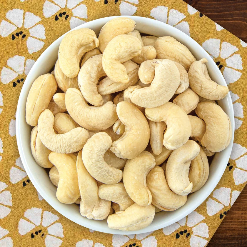 A small white bowl full of organic raw cashes, sitting on a yellow cloth with a floral pattern, on top of a dark brown wooden surface