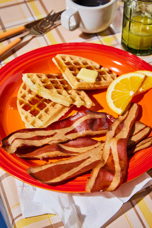 A red breakfast plate with a waffles, slices of oranges, and several pieces of Prime Roots Applewood Smoked Koji Bacon