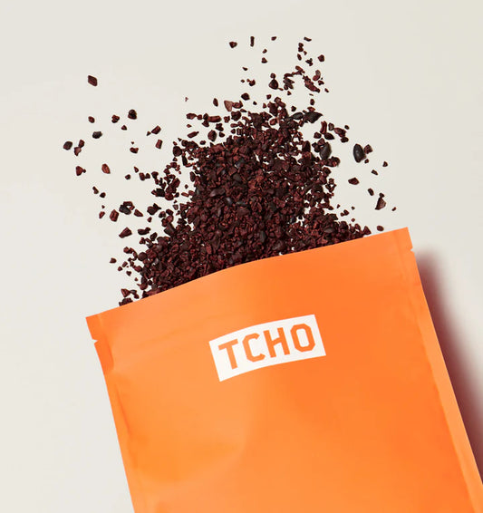 An orange pouch with the brand name 'TCHO' printed in white, shown with dark brown crushed cacao nibs spilling out against a light neutral background.
