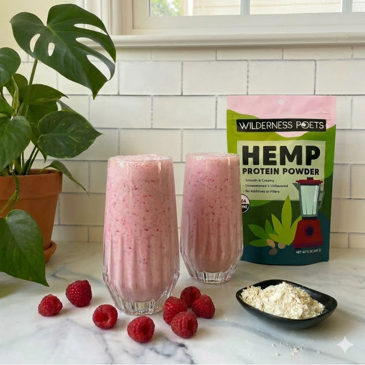 Two glasses of pink smoothie with raspberries, a bag of Wilderness Poets Hemp Protein Powder, and a bowl of protein powder on a marble countertop.
