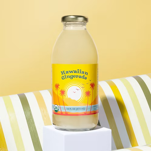 A bottle of Big Island Organics Hawaiian Gingerade beverage with a yellow tropical sunset label, standing on a white pedestal against a yellow background with striped fabrioc.