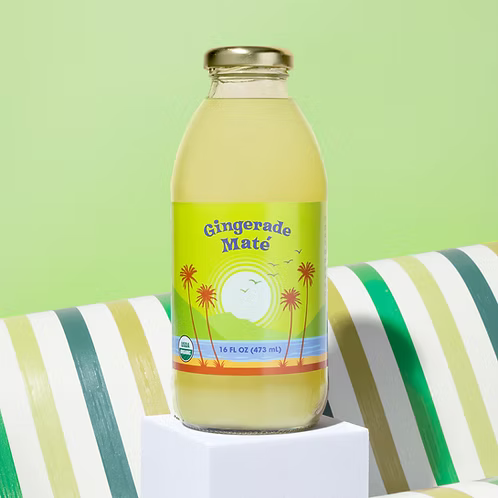 A bottle of Big Island Organics Gingerade Mate iced tea sits on a white pedestal. The clear glass bottle shows the pale yellow liquid inside and has a colorful label with palm trees and a sun.