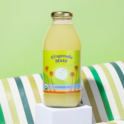 A bottle of Big Island Organics Gingerade Mate iced tea sits on a white pedestal. The clear glass bottle shows the pale yellow liquid inside and has a colorful label with palm trees and a sun.