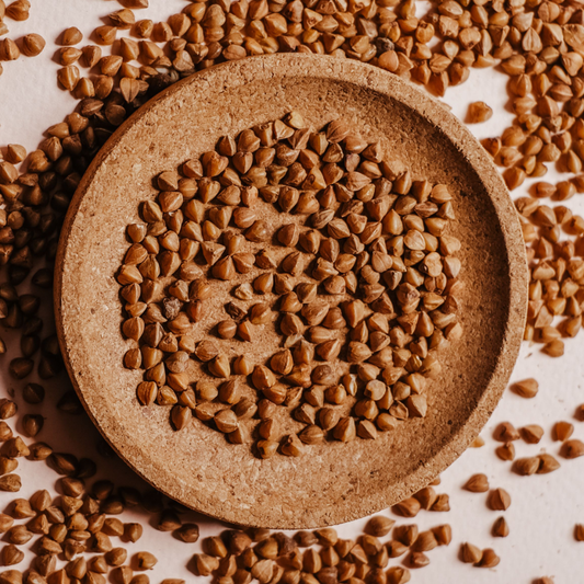 A top-down view of brown buckwheat groats arranged in a round beige ceramic dish, with additional scattered buckwheat seeds around the dish on a light background.