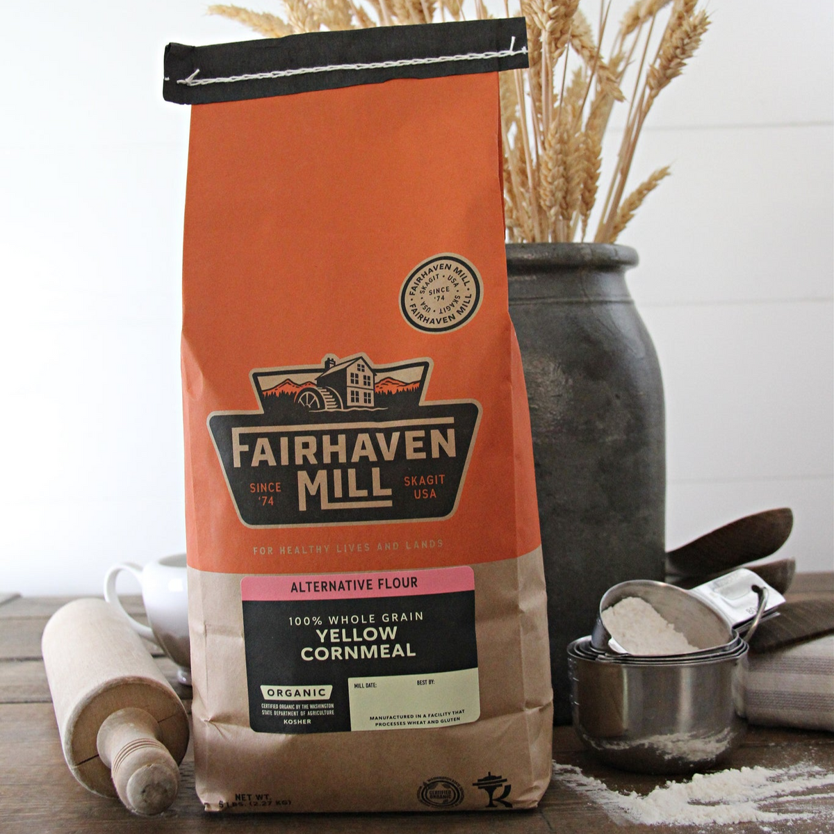 A 25 lb bulk bag of Fairhaven Mill Organic Yellow Cornmeal, with a rolling pin and a measuring cup with cornmeal in the foreground.