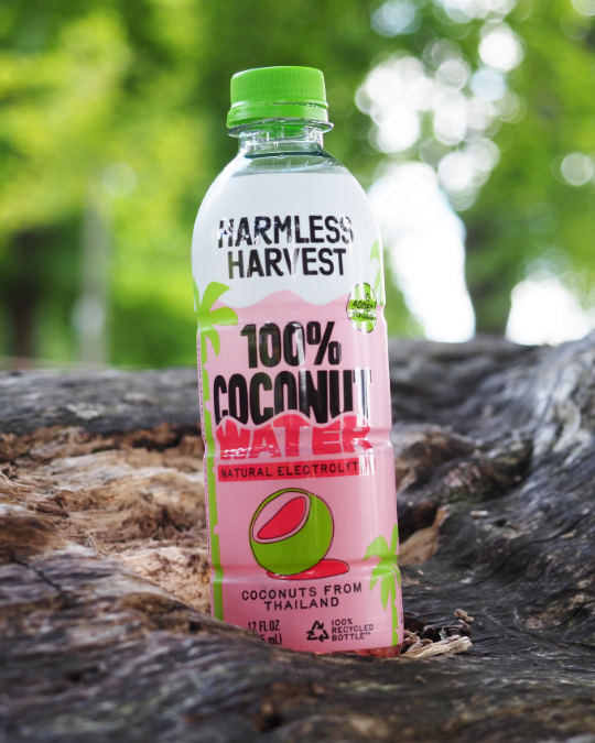 A bottle of Harmless Harvest 100% Coconut Water, natural electrolyte drink, standing on a rustic log with a blurred green forest background.