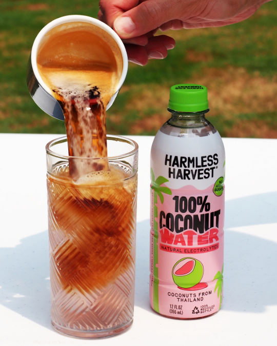 A bottle of Harmless Harvest 100% Coconut Water is shown next to a glass of coconut water with coffee being poured into it, set on a white table with a blurred green grass background.