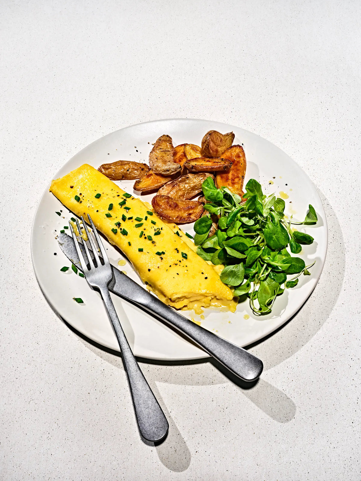 A plated breakfast featuring a folded Just Egg omlette with herbs, roasted potato wedges, and green salad on a white plate with a fork and knife, set on a light speckled countertop.