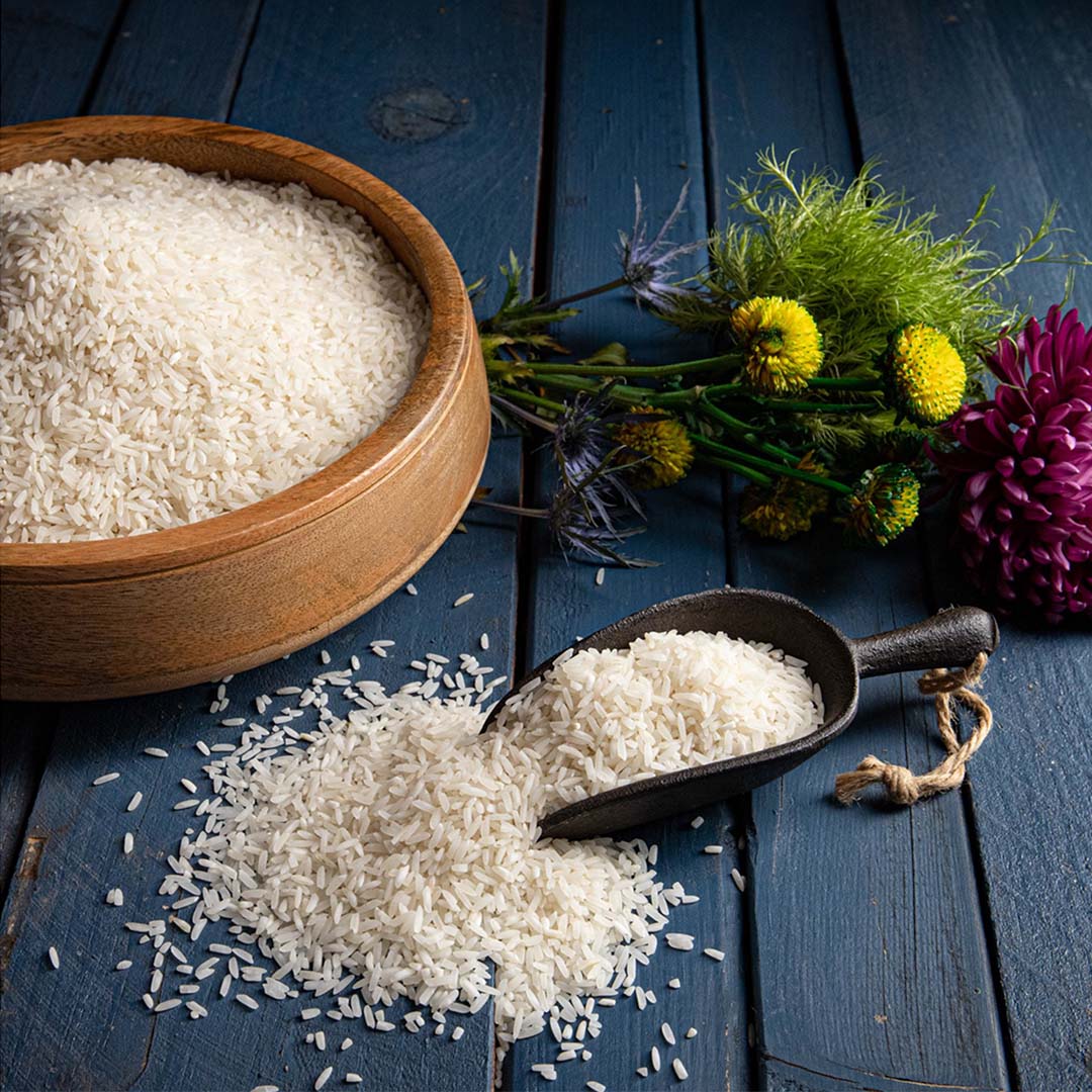 An iron scoop overflowing with white basmati rice, next to a large wooden bowl filled with white basmati rice, in front of some picked flowers laying on a blue painted wooden surface