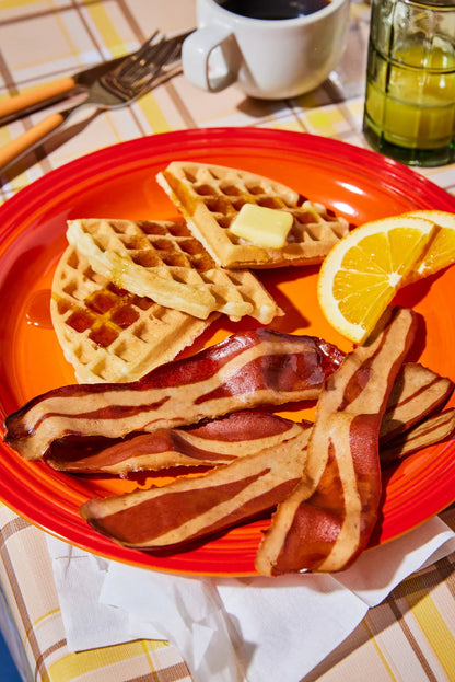 A red breakfast plate with a waffles, slices of oranges, and several pieces of Prime Roots Applewood Smoked Koji Bacon