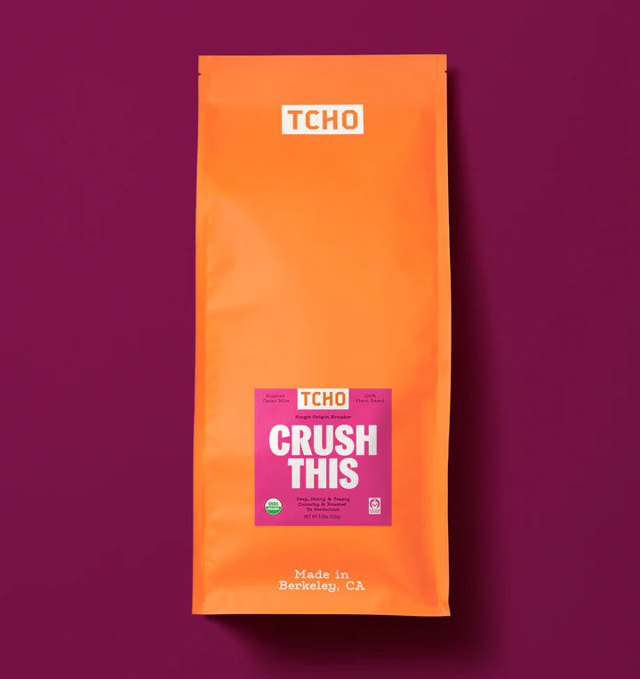 An orange stand-up pouch of TCHO Crush This cacao nibs on a solid dark magenta background. The package features a white TCHO logo at the top and a pink and white 'CRUSH THIS' label in the center.
