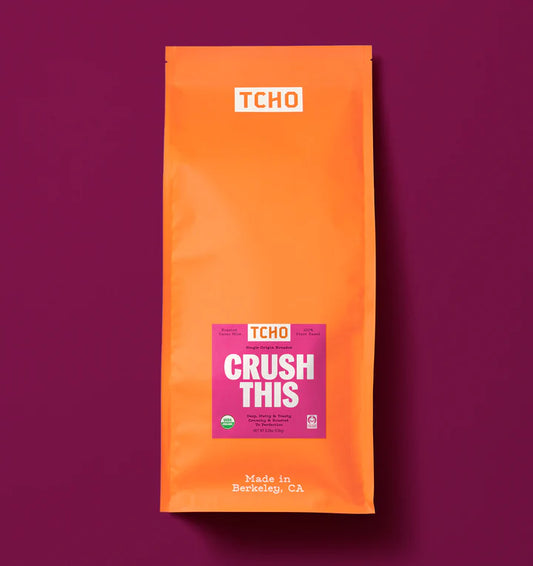 An orange stand-up pouch of TCHO Crush This cacao nibs on a solid dark magenta background. The package features a white TCHO logo at the top and a pink and white 'CRUSH THIS' label in the center.