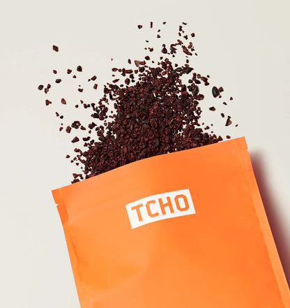 An orange pouch with the brand name 'TCHO' printed in white, shown with dark brown crushed cacao nibs spilling out against a light neutral background.